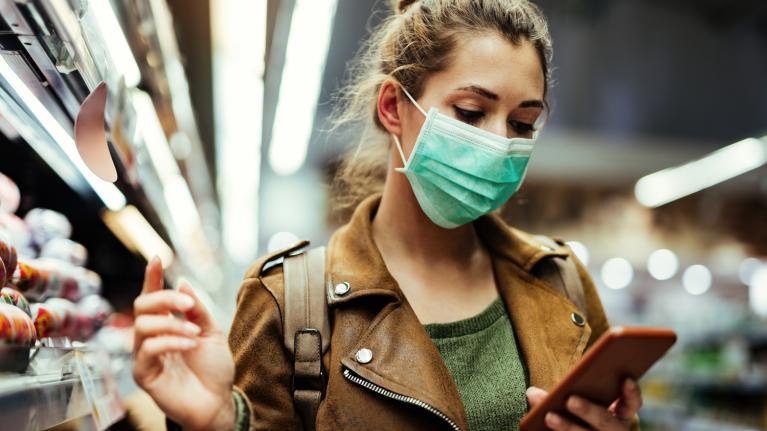 young woman with face mask at grocery store looking at her phone