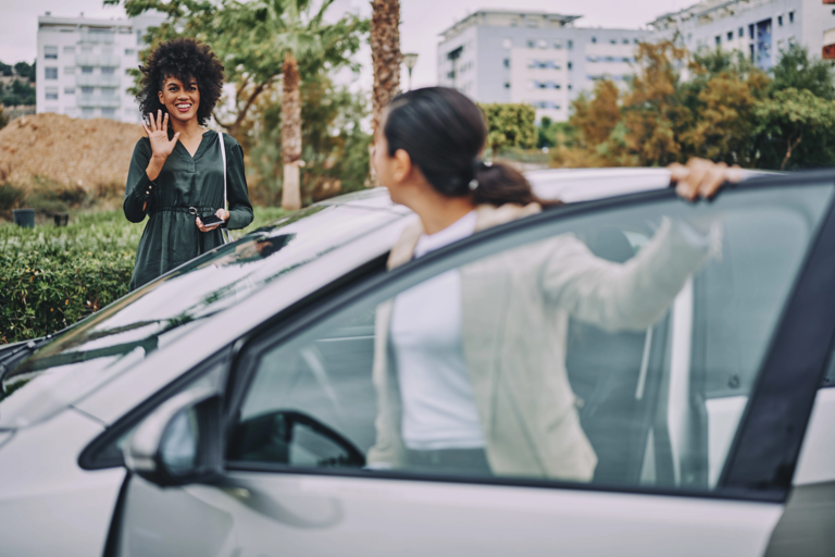 A woman waves at her friend who is getting out of her car.